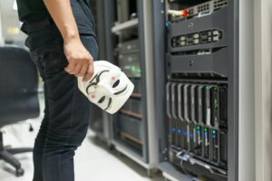 Une personne tenant un masque anonyme près des serveurs, faisant allusion à des thèmes de cybersécurité et de piratage.